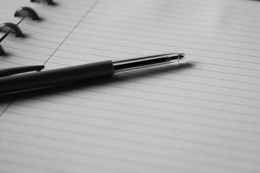 Home Black and white image of a pen resting on an open lined notebook, ideal for writing or note-taking themes.